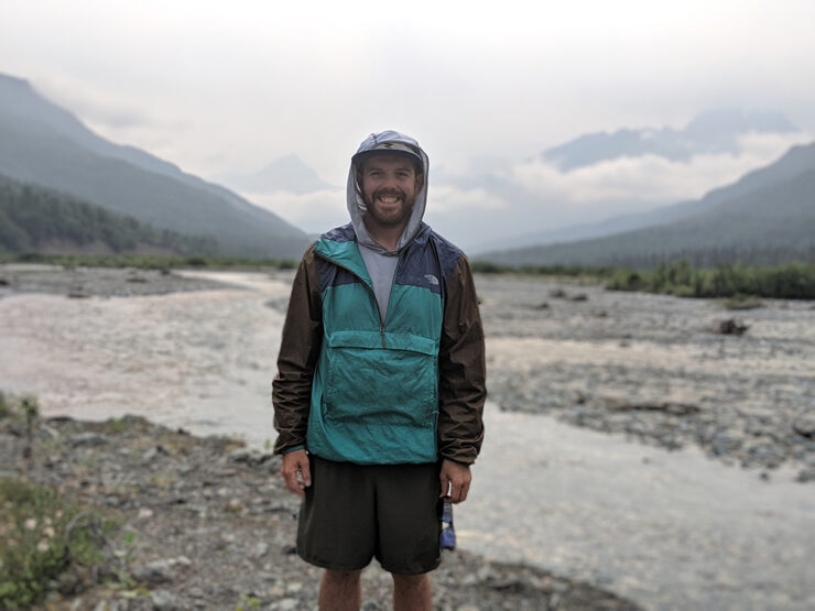 James DiRosa standing in shorts and a parka in front of a mountain snowy mountain stream