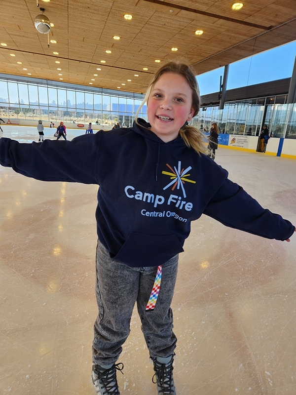 A camper ice skating with arms outstretched and wearing a navy blue Camp Fire sweatshirt.