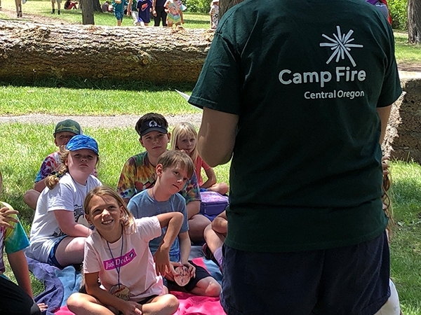 A counselor wearing a Camp Fire t-shirt and talking to campers sitting in the grass.