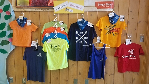 An assortment of colorful Camp Fire t-shirts hanging in a display on a pine wood wall.