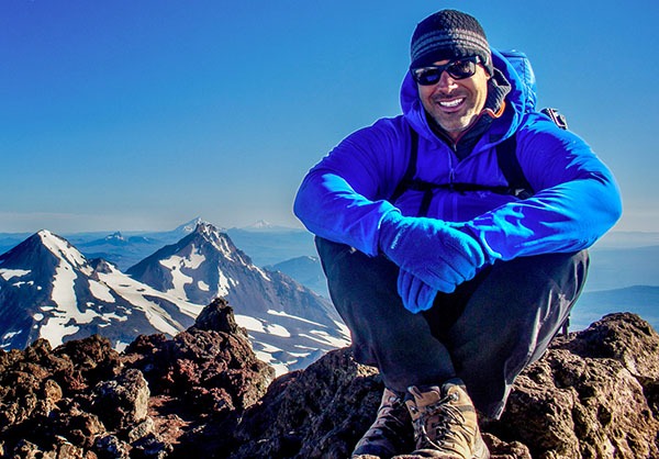 Joey Hamilton sitting on the high rocky summit of South Sister surrounded by snowy peaks.