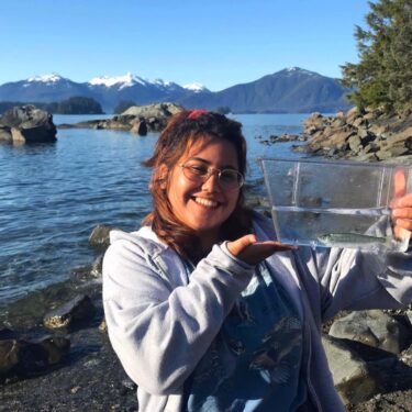 Picture of Cyprianna Martinez holding a fish in a container in front of a body of water.